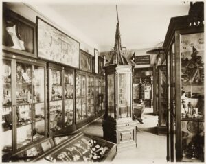 Sepiowe zdjęcie sali muzealnej wypełnionej szklanymi gablotami zawierającymi różne artefakty i przedmioty. Pośrodku stoi wysoka, ozdobna gablota. Gabloty ścienne z napisami "Japonia" i "Chiny" prezentują skomplikowane przedmioty, a nad gablotami wiszą oprawione dzieła sztuki.