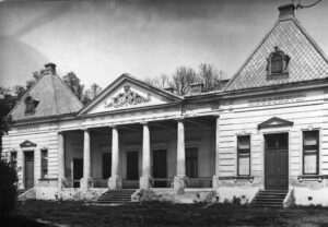 Czarno-białe zdjęcie neoklasycystycznego budynku z centralnym portykiem wspartym na sześciu kolumnach i dwoma skrzydłami po obu stronach. Dach jest stromy z trójkątnymi frontonami. Niektóre okna i drzwi są zamknięte lub zabite deskami. W tle widać drzewa.