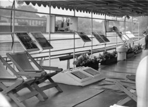 Czarno-białe zdjęcie zadaszonego pokładu łodzi z pustymi drewnianymi leżakami, skośnym otworem wentylacyjnym, oprawionymi zdjęciami na stojakach, skrzynkami na kwiaty i latarniami. Łódź jest zadokowana w pobliżu wody, a w tle widoczne są budynki i drzewa.